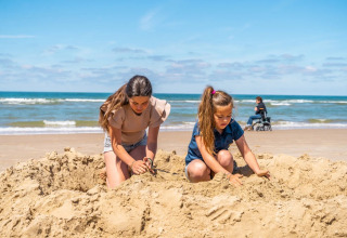 To piger leger i sandet på en strand ved en feriepark med glamping, kvinde i kørestol bagved.