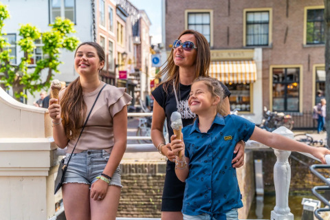 Gezin geniet van een ijsje in de zon bij een vakantiepark met glamping en charmante gebouwen daarachter.
