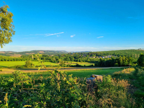 Vakantiepark Camping & Ferienpark Brilon met groene weilanden, bossen en een dorpje in de verte onder blauwe lucht.