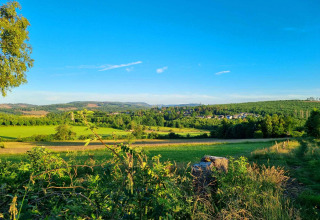 Vakantiepark Camping & Ferienpark Brilon met groene weilanden, bossen en een dorpje in de verte onder blauwe lucht.