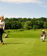 Two golfers on a green course near Brilon, Germany, with lush trees and golf bags visible in the background.