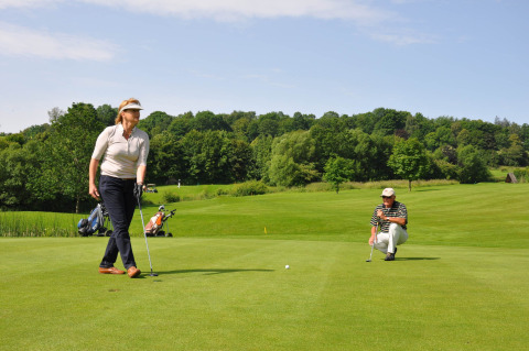 Two golfers on a green course near Brilon, Germany, with lush trees and golf bags visible in the background.