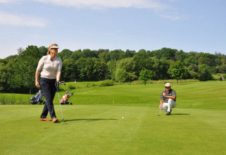Zwei Golfspieler auf einem Rasenplatz bei Brilon, Deutschland, mit Bäumen und Golftaschen im Hintergrund.