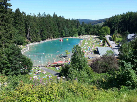 View of swimming lake and sunbathers at Camping & Ferienpark Brilon, surrounded by forest in Germany.