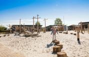Barn leger på legeplads med træstammer og sand på en feriepark med glampingboliger og blå himmel.