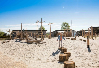 Barn leger på legeplads med træstammer og sand på en feriepark med glampingboliger og blå himmel.
