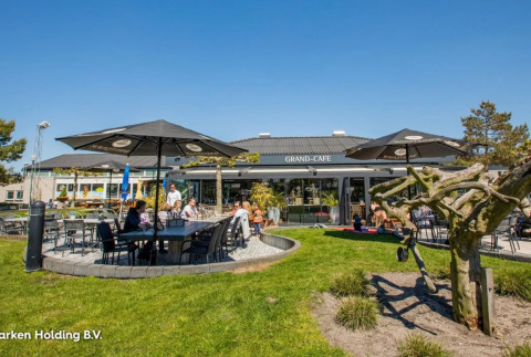 Área de cafetería al aire libre en un parque vacacional con mesas, sombrillas y familias disfrutando el glamping.
