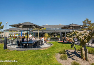 Buitencafé bij een vakantiepark met tafels, parasols en families die genieten van glamping faciliteiten.