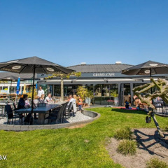 Area caffè all'aperto in un parco vacanze con tavoli, ombrelloni e famiglie che godono del glamping.