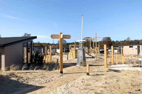 Holiday park with glamping cabins, sandy grounds, playground, and signpost pointing 'Zuid' and 'Noord'.