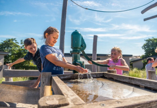 Børn leger med en vandpumpe på en udendørs legeplads i solskinsvejr på en feriepark med glamping.