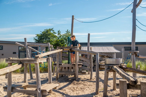 Kind speelt op een houten speeltoestel bij een vakantiepark met glamping onder een heldere zonnige lucht.