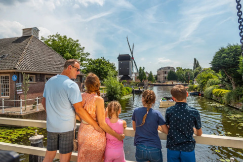 Gezin op brug kijkt uit over een kanaal en molen bij een vakantiepark met glamping accommodaties.