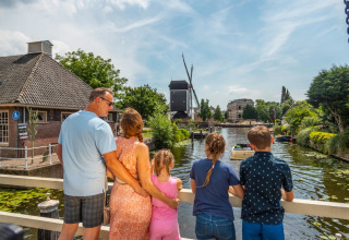 Family stands on a bridge overlooking a canal and windmill at a holiday park offering glamping stays.