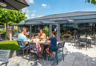 Familie nyder udendørs måltid ved et bord foran Grand Café på en feriepark med glamping faciliteter.