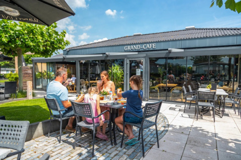 Familie nyder udendørs måltid ved et bord foran Grand Café på en feriepark med glamping faciliteter.