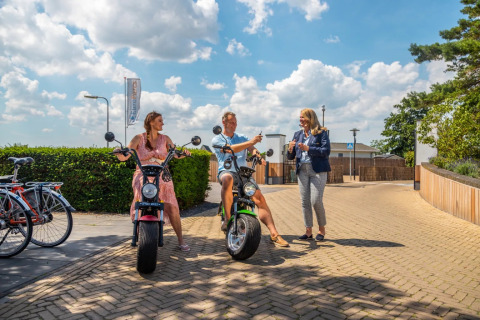 Huéspedes disfrutan scooters eléctricos en un parque vacacional con alojamiento glamping bajo cielo soleado.