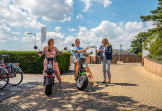 Gæster nyder el-scootere og solskin ved en feriepark med glamping-indkvartering under blå himmel.