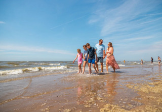 Familie går langs en strand ved et feriepark med glamping, under en klar blå himmel og roligt hav.