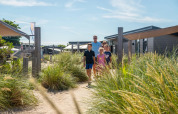 Familie går på sti blandt græs ved moderne glamping-indkvartering i en feriepark under blå himmel.
