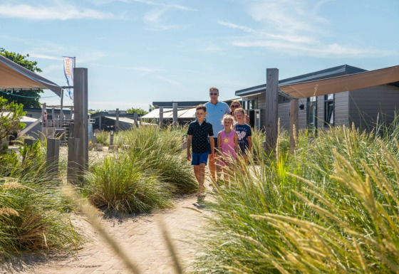 Familie går på sti blandt græs ved moderne glamping-indkvartering i en feriepark under blå himmel.