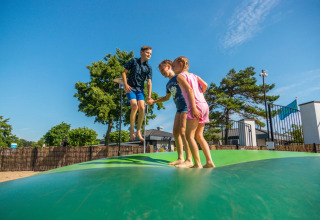 Drie kinderen springen op een luchtkussen in een vakantiepark met zonnige glamping accommodaties.