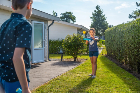 Dos niños juegan con palas y pelota frente a su alojamiento glamping en un parque vacacional soleado.