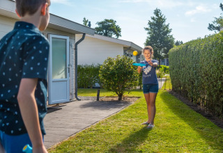 To børn leger bat og bold udenfor ved deres glamping-indkvartering i en feriepark på en solrig dag.