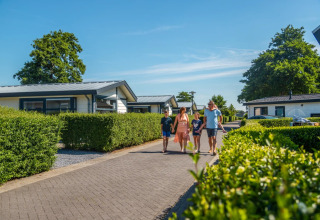 Familie wandelt in een vakantiepark met glampingverblijven tussen groene hagen onder een blauwe lucht.