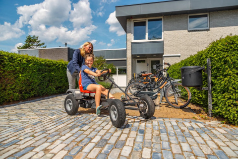 Zwei Personen genießen eine Fahrt mit einem Kettcar vor einem modernen Ferienhaus in einem Ferienpark.