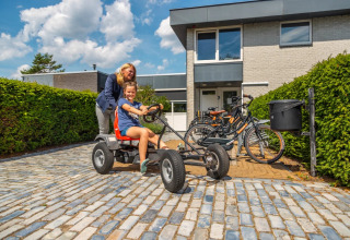 Two people enjoy a go-kart ride in front of a modern holiday park accommodation with bikes and green hedges.