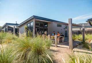 Family relaxing outside a modern glamping cabin at a holiday park, surrounded by grass and blue sky.