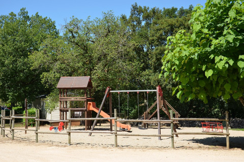 Parque infantil con columpios y tobogán en Camping le Roc, rodeado de árboles en Occitanie, Francia.