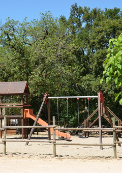 Parque infantil con columpios y tobogán en Camping le Roc, rodeado de árboles en Occitanie, Francia.