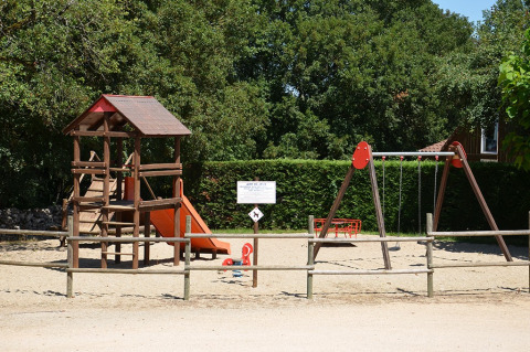 Parque infantil con juego de trepar, tobogán y columpios en Camping le Roc, Occitania, Francia, rodeado de árboles.