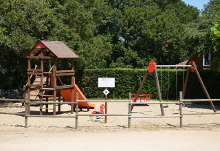 Parque infantil con juego de trepar, tobogán y columpios en Camping le Roc, Occitania, Francia, rodeado de árboles.