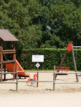 Parque infantil con juego de trepar, tobogán y columpios en Camping le Roc, Occitania, Francia, rodeado de árboles.