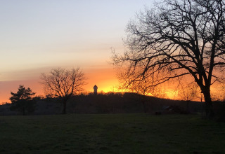 Tramonto al Camping le Roc, Occitanie, Francia, con sagome di alberi e una torre dell'acqua in lontananza.