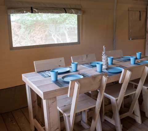 Interno di una tenda safari con tavolo in legno, sei sedie e stoviglie blu, apparecchiato per sei persone.