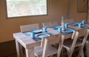 Intérieur d’une tente safari avec table en bois, six chaises et vaisselle bleue, prête pour un repas.
