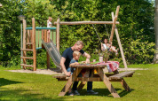 Family enjoys a picnic outdoors while children play on a wooden playground set surrounded by greenery.