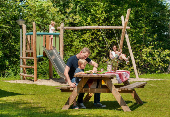 Family enjoys a picnic outdoors while children play on a wooden playground set surrounded by greenery.
