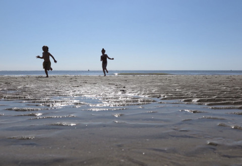 Two children run and play on a sandy beach at low tide under a clear blue sky in Zeeland, Netherlands.
