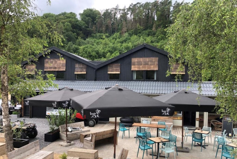 Zona de cafetería al aire libre en Camping De La Vallée, Bélgica, rodeada de vegetación y edificio moderno.