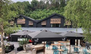 Zona de cafetería al aire libre en Camping De La Vallée, Bélgica, rodeada de vegetación y edificio moderno.