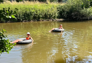 Two people relax on inflatable rings floating down a calm river, surrounded by lush greenery and trees.