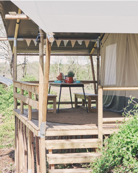 Houten terras met zitbank en tafel aan een safaritent, versierd met vlaggetjes, midden in de natuur.