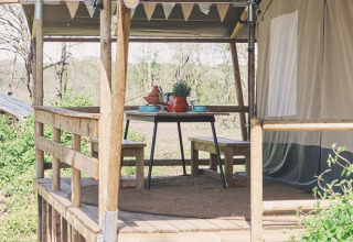 Terrasse en bois avec table à manger devant une tente safari, décorée de fanions et entourée de nature.