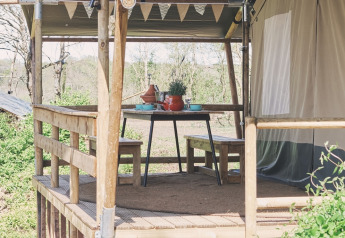 Veranda in legno con tavolo e panche da pranzo davanti a una tenda safari immersa nella natura.