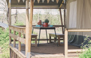 Wooden deck with dining table and benches outside a safari tent, decorated with bunting in nature.
